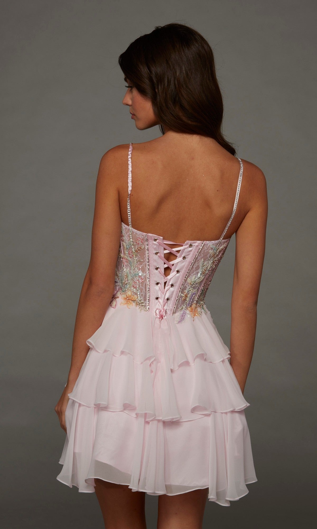 A woman is shown from behind wearing the Alyce 30096 short homecoming dress in light pink, featuring a sleeveless fit-and-flare silhouette, lace-up back, floral embroidery, and a layered ruffle skirt against a plain background.